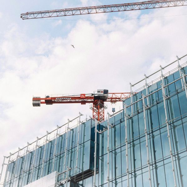 Two large construction cranes, one red, hover above a modern glass building façade against a cloudy blue sky.