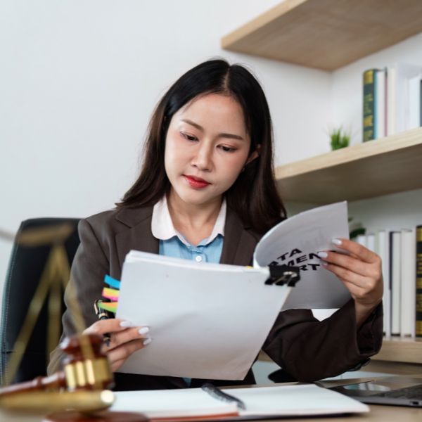 A legal administrator reviews documents at her desk with a small gavel in the foreground and bookshelves behind her.