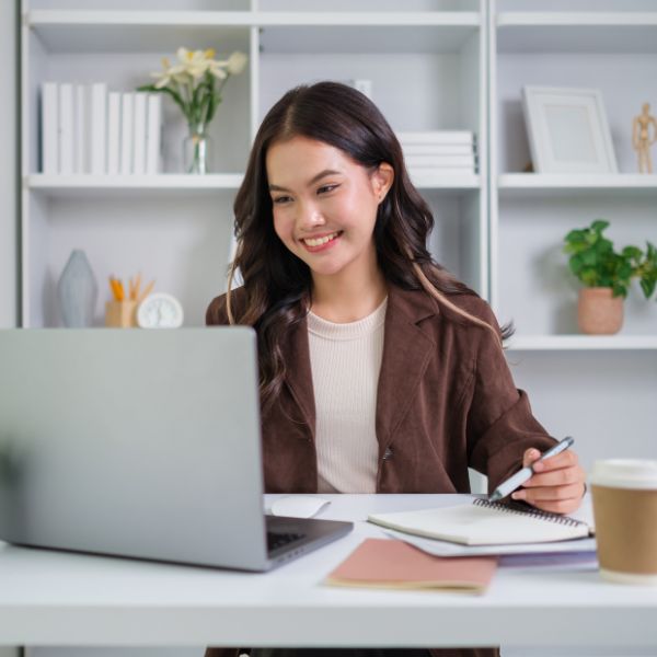 A smiling young woman works at her laptop, taking notes at a desk in front of a bright, white bookshelf.