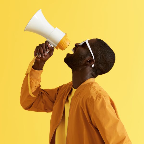 A man in an orange jacket and white sunglasses shouts into a white and yellow megaphone against a yellow background.